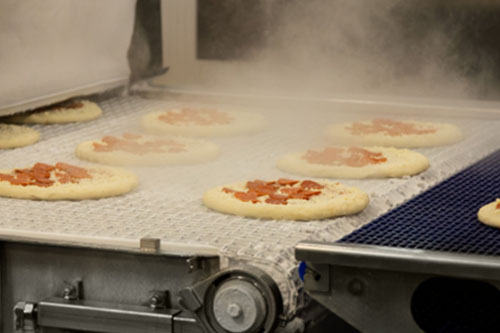 Industrial food production line where pizzas with toppings move along a metal conveyor, with visible steam, in an automated processing environment.